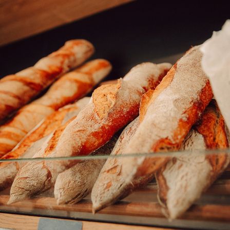 Eine Nahaufnahme von frisch gebackenen Baguettes auf einem Glasregal, mit einem Holzboden und etwas mit Mehl bestäubt.
