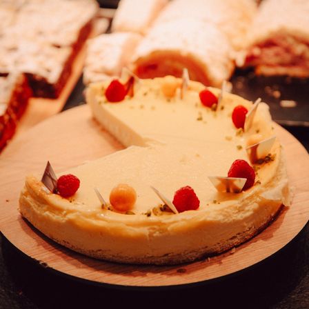 Ein Stück Käsekuchen mit cremiger Füllung, verziert mit Himbeeren und Pistazien, steht auf einem Holzteller. Im Hintergrund sind weitere Desserts zu sehen, darunter eine Rolle und ein Kuchen.