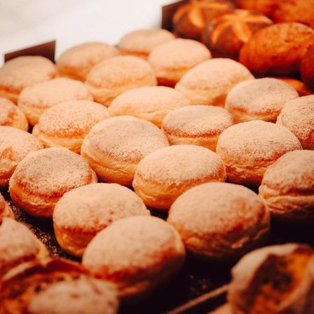Ein Nahaufnahme von einem Tablett gefüllt mit frisch gebackenen, puderzuckerbedeckten Donuts. Die Donuts sind in Reihen angeordnet, und einige sind etwas größer als andere. Das Tablett ist dunkel, möglicherweise aus Holz.