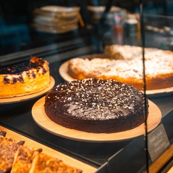 Eine Ausstellung verschiedener Desserts, darunter ein Schokoladenkuchen mit Streuseln, ein Blaubeerkuchen und ein Sahnekuchen, alles hinter einer Glasvitrine.