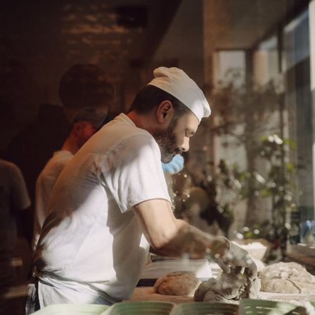 Ein Mann in einer weißen Kochmütze knetet Teig auf einem Tisch mit anderem Teig und Brot.