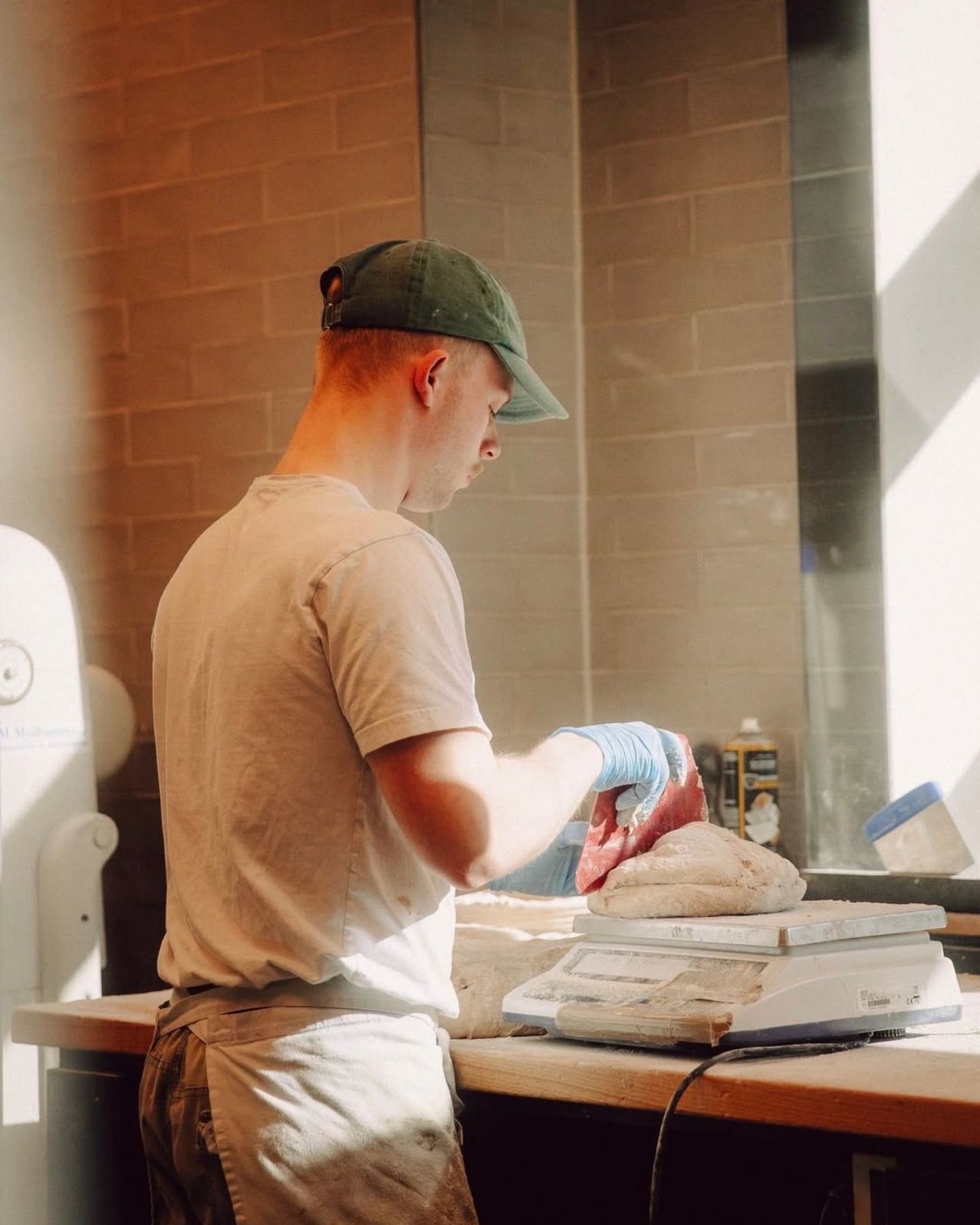 A man wearing a cap and gloves works in a kitchen, handling dough on a scale.