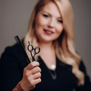 Eine Frau mit blonden Haaren und Tattoos hält Schere und Kamm und lächelt für ein Foto. Sie trägt ein schwarzes Oberteil.