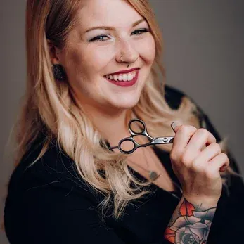 Eine Frau mit blondem Haar hält eine Schere in der Hand und lächelt für ein Foto. Sie hat Tattoos am Arm und einen Ring in der Nase.