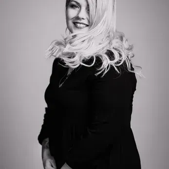 Ein schwarz-weißes Porträt einer lächelnden Frau mit langen, welligen blonden Haaren, die ein schwarzes Oberteil trägt, steht und für ein Foto posiert.