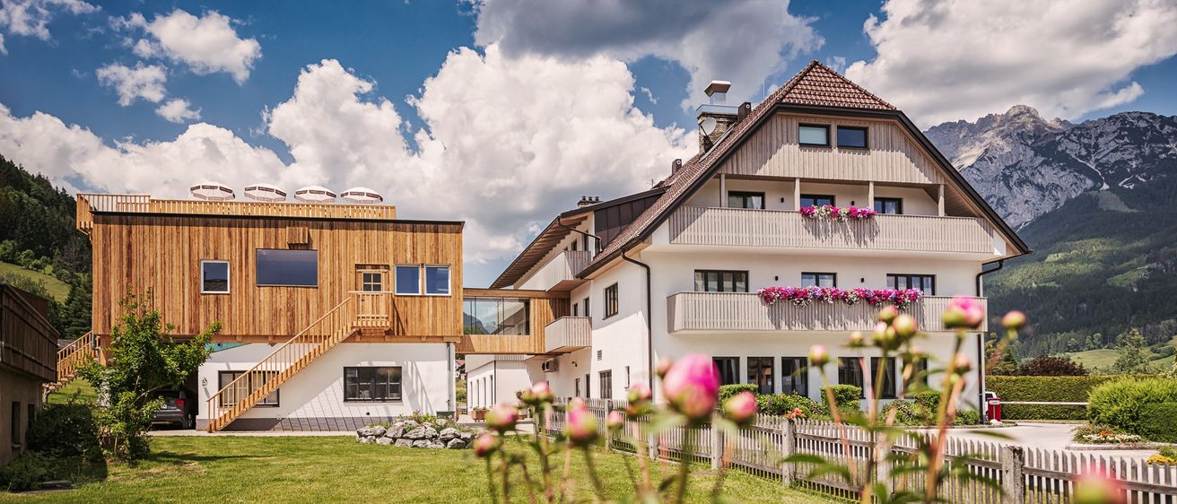 Zwei moderne Häuser mit Balkonen und Blumen stehen unter einem teilweise bewölkten Himmel. Die Häuser sind von einem Rasen und Blumen umgeben.