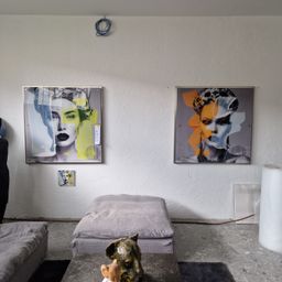 Ein Raum mit zwei abstrakten Gemälden an der Wand. Das linke Gemälde zeigt eine Frau mit einem gelben und weißen Gesicht, und das rechte Gemälde zeigt eine Frau mit einem orangefarbenen Gesicht. Ein grauer Hocker und eine Skulptur stehen auf dem Boden.