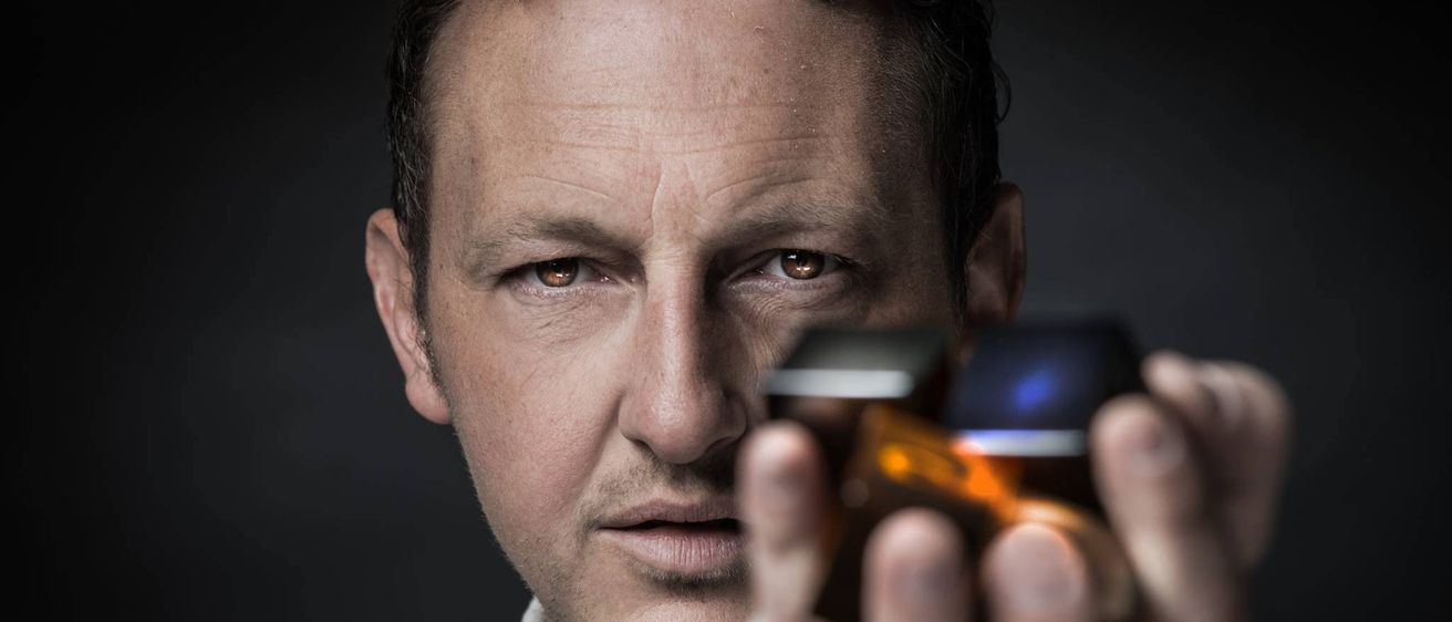 Ein Mann mit braunen Augen hält ein kleines elektronisches Gerät in den Händen. Er scheint es genau zu untersuchen.