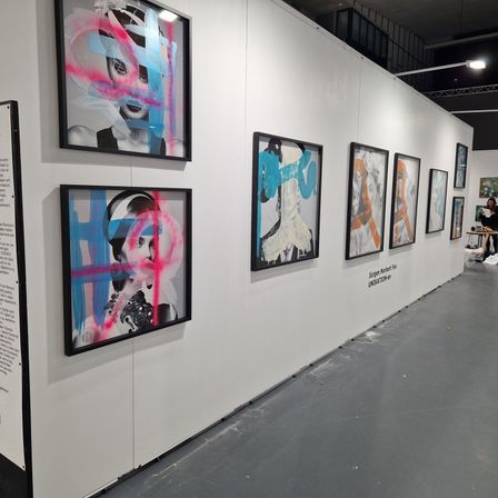 Eine Ausstellungswand zeigt mehrere abstrakte Gemälde von Frauen in verschiedenen Farben und Stilen. Eine Person sitzt in der Ecke.