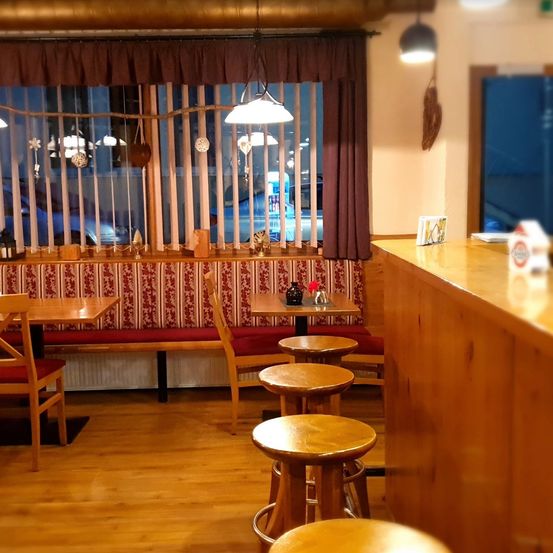 Ein Restaurant-Innenraum mit Holzböden und einer hölzernen Theke. Es gibt Tische und Stühle, eine Bank am Fenster und eine Lampe. Draußen ist ein Auto durch das Fenster sichtbar.