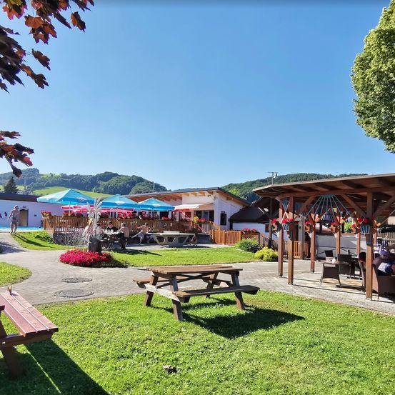 Freiluft-Essbereich mit Picknicktischen und Bänken. Sonnenschirme und Blumen vor einem Restaurant. Bäume und Berge im Hintergrund.