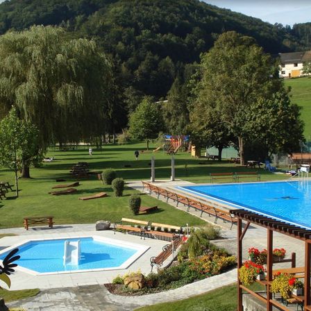 Ein Blick von oben auf einen Ferienort mit zwei Pools, einem großen und einem kleinen, umgeben von grünen Rasenflächen, Bäumen und einem Spielplatz. Das Gebiet verfügt auch über Bänke, Tische und ein Haus in der Ferne.