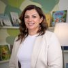 Eine Frau mit braunen Haaren lächelt in die Kamera. Sie trägt ein weißes Hemd und einen beigen Blazer. Sie steht vor einem Bücherregal mit Büchern und einer Lampe.