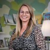 Eine Frau in einem Leopardenmusterkleid steht lächelnd in einem Raum mit Bücherregalen und einer Lampe, umgeben von Kinderbüchern.