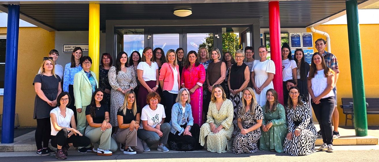 Eine Gruppe von Frauen posiert vor dem 'Volksschule'-Gebäude, lächelt und schaut in die Kamera. Das Schild darüber ist bunt und steht 'Volksschule'.