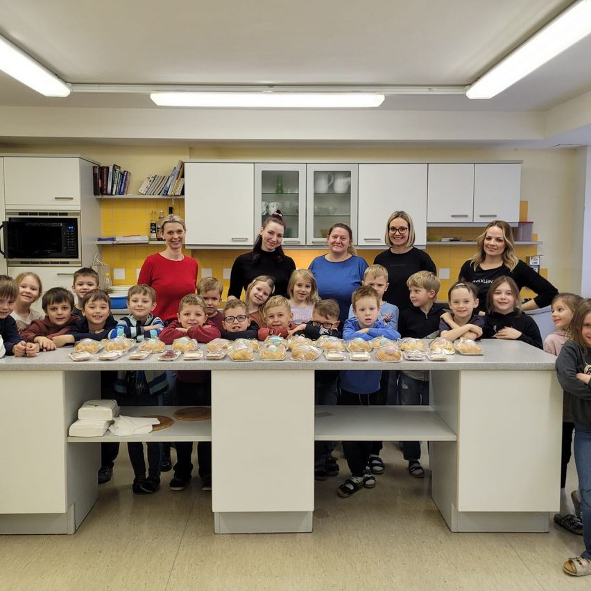 Eine Gruppe von Kindern und Erwachsenen steht hinter einem Tisch mit Essen. Einige Kinder tragen Brillen. Im Hintergrund befinden sich Schränke und eine Mikrowelle.