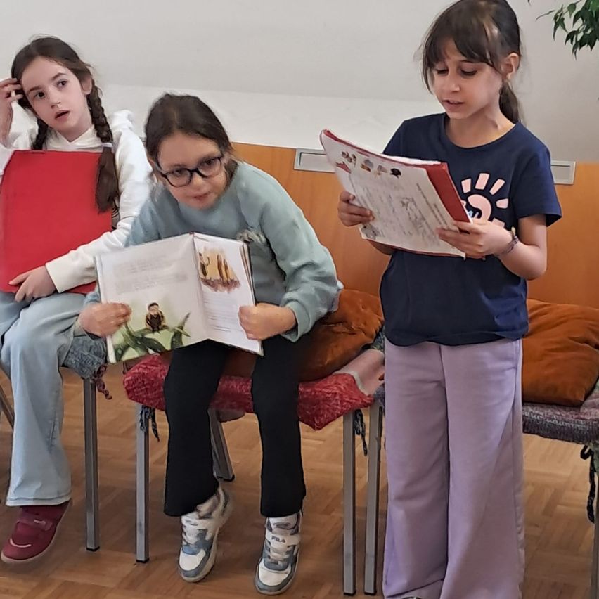 Drei Mädchen sitzen auf Stühlen. Ein Mädchen liest ein Buch, ein anderes Mädchen liest einen roten Ordner und das dritte Mädchen schaut sich das Buch an.