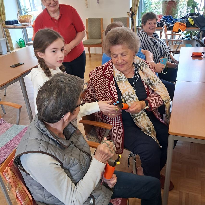 In einem Raum sind ältere Frauen versammelt, eine bastelt mit Werkzeugen, während eine andere ein Baby hält. Eine Frau in Rot steht dahinter und andere sind sitzend.