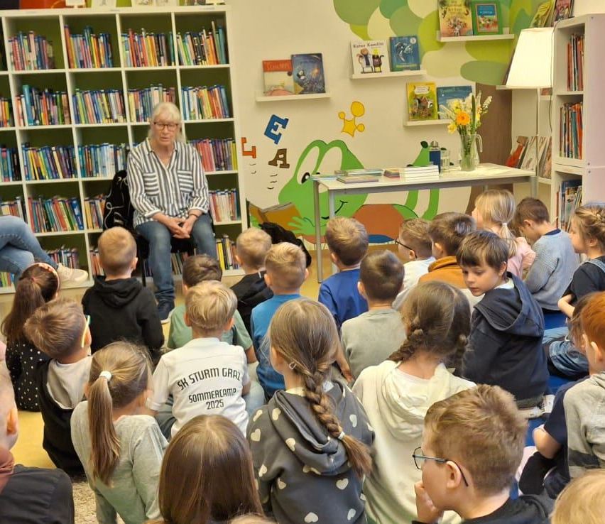 Eine Frau liest Kindern in einer Bibliothek ein Buch vor. Die Kinder sitzen auf dem Boden und lauschen aufmerksam.