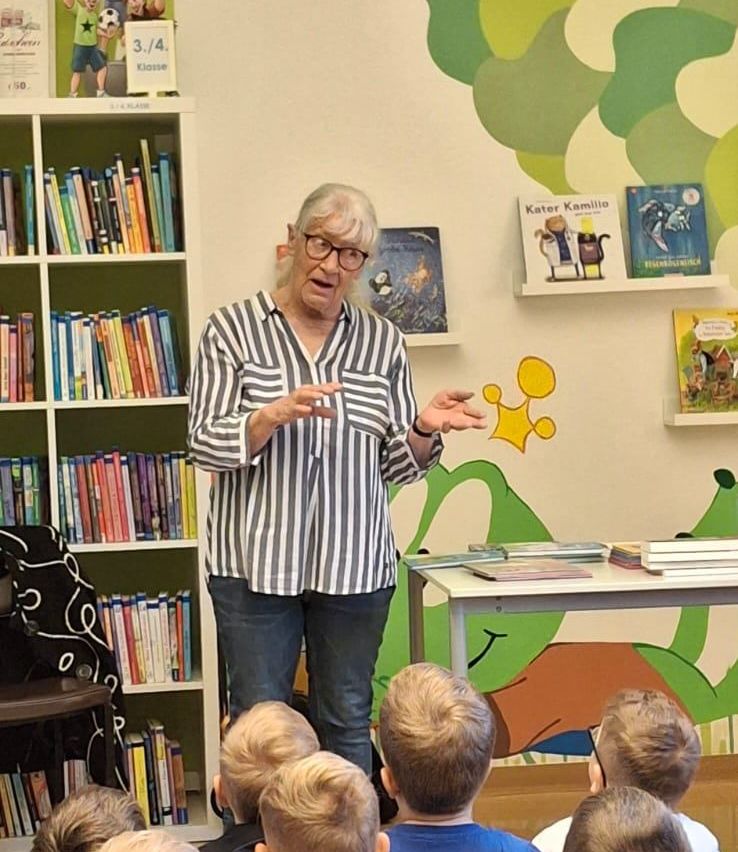 Eine ältere Frau mit Brille und einem gestreiften Shirt steht vor Kindern in einem Klassenzimmer. Bücher stehen hinter ihr auf Regalen und auf dem Tisch.