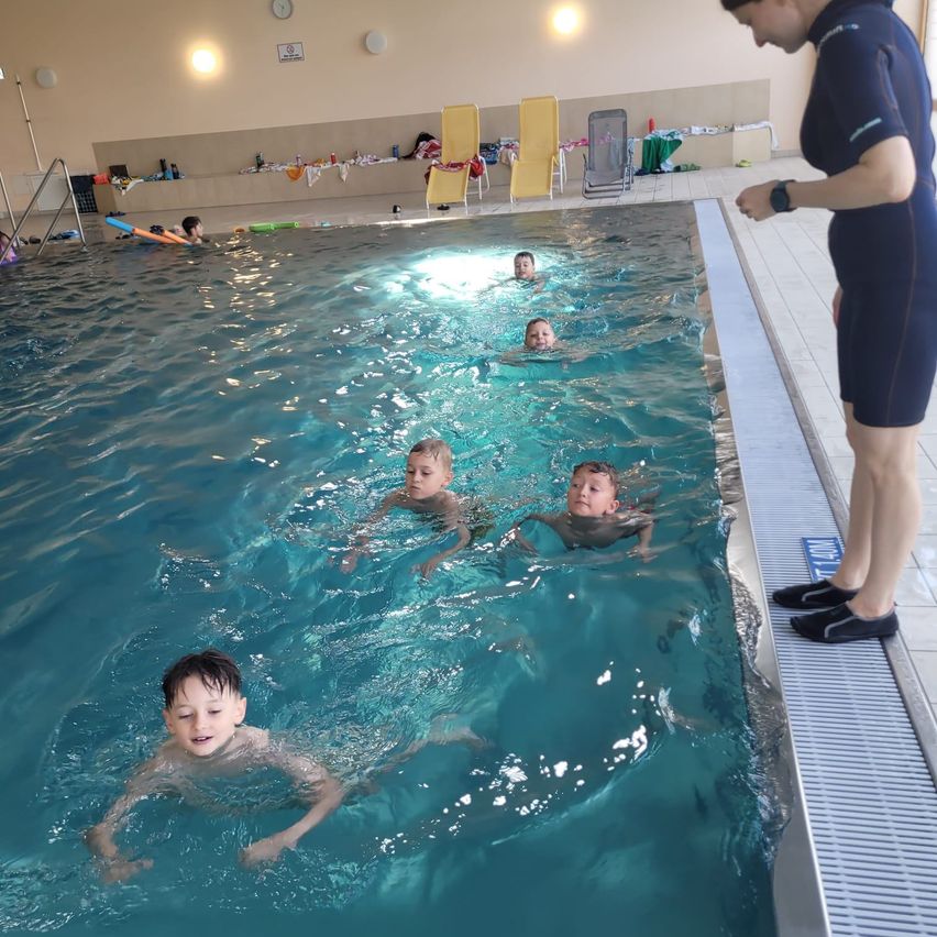 Eine Schwimmlehrerin unterrichtet Kinder im Schwimmen in einem Pool. Einige Kinder schwimmen, während andere auf Schwimmhilfen treiben.