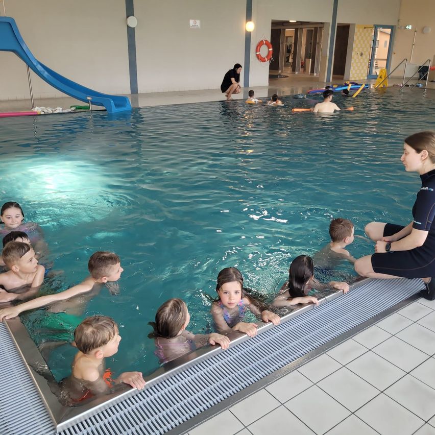 Mehrere Kinder schwimmen in einem Hallenbad, mit einem Rettungsschwimmer an der Seite. Eine Rutsche und ein Rettungsring sind sichtbar.
