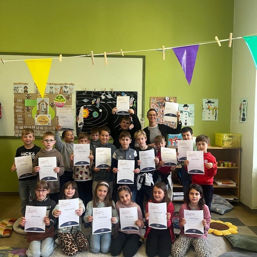 Ein Klassenzimmer voller Kinder, die Urkunden halten und für ein Foto posieren, mit zwei Lehrern hinter ihnen. Sie befinden sich in einem Raum mit grünen Wänden, bunten Wimpelketten und einer Bulletin-Board.
