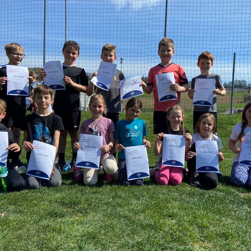 Eine Gruppe von Kindern und Jugendlichen posiert für ein Foto auf einem Rasenfeld, während sie Urkunden halten. Der Hintergrund zeigt einen Netzzaun und ein Feld.