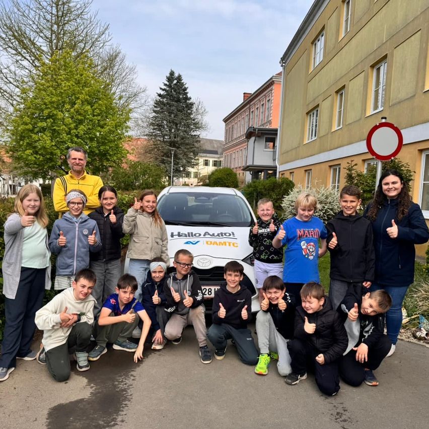 Eine Gruppe von Kindern und Erwachsenen posiert vor einem Toyota-Auto mit dem Nummernschild 28, lächelt und gibt Daumen hoch. Sie sind draußen in einem Vorstadt-Setting.