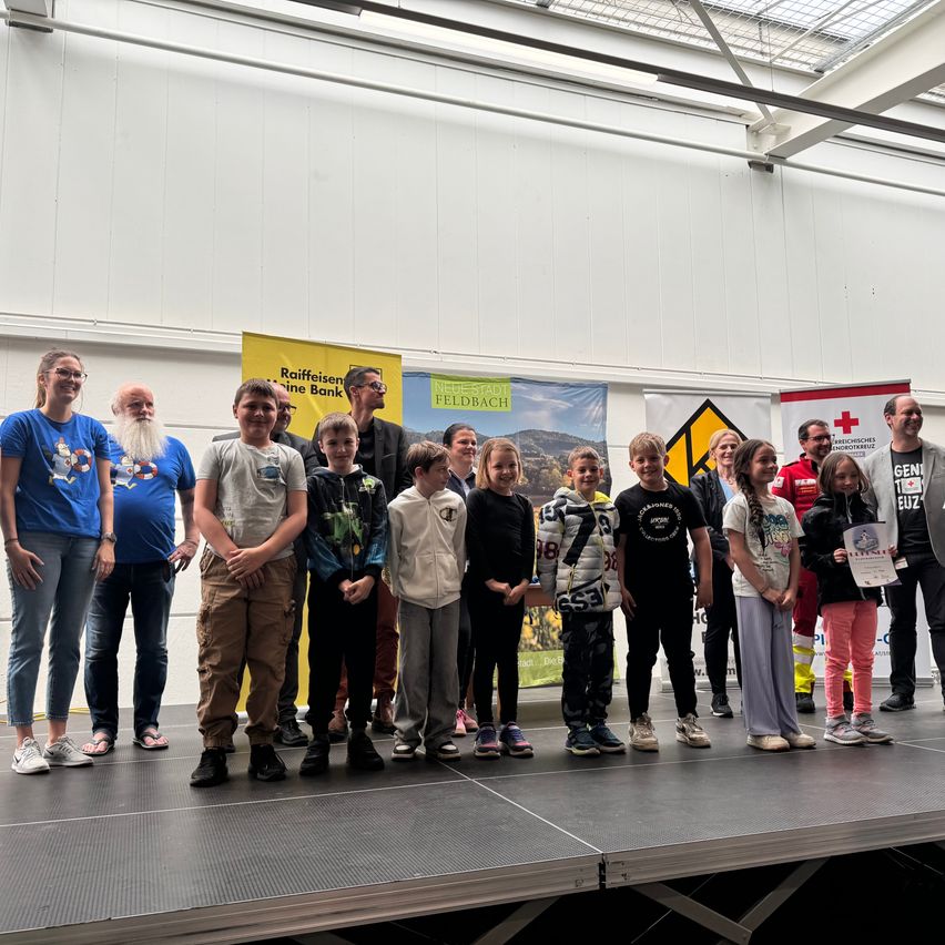 Gruppe von Kindern und Erwachsenen steht auf einer Bühne, einige halten Urkunden. Dahinter sind Banner und Schilder angebracht.