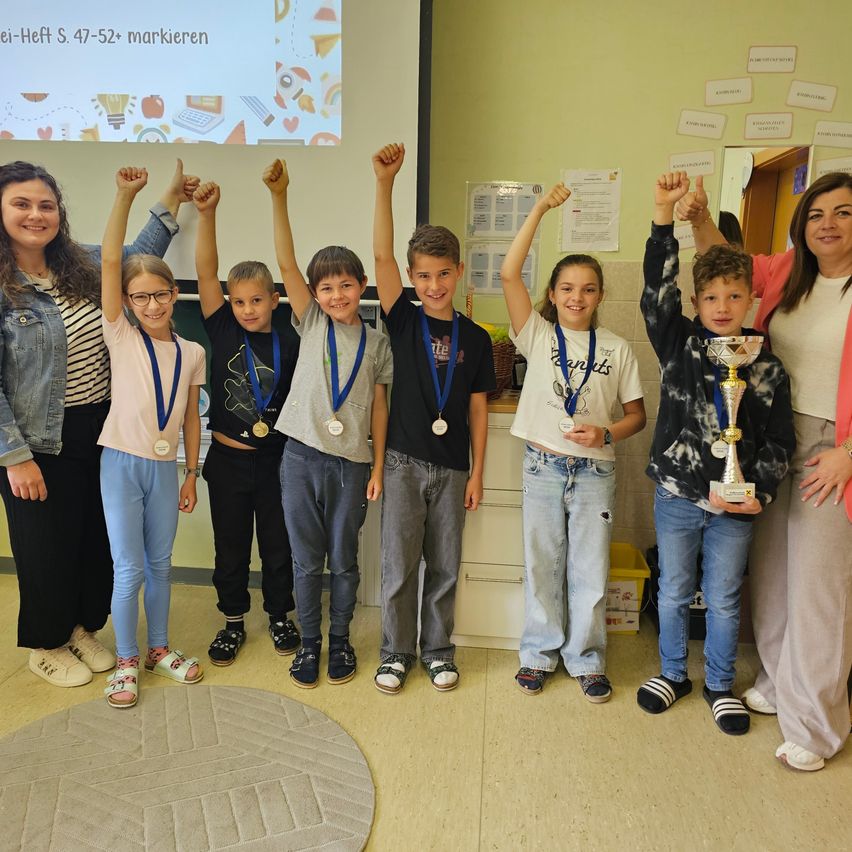 Eine Gruppe von Kindern mit Medaillen um den Hals feiert in einem Klassenzimmer mit zwei lächelnden Frauen.