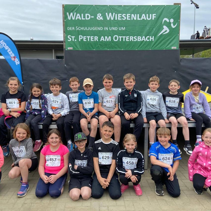 Eine Gruppe von Kindern mit Nummern auf ihren Trikots posiert für ein Foto mit einem Banner hinter ihnen, das Wald- & Wiesenlauf steht.