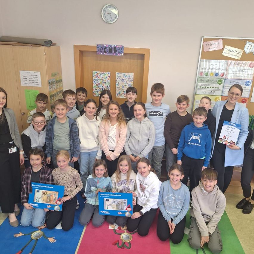 Eine Gruppe von Kindern und Lehrern posiert für ein Foto im Klassenzimmer. Einige Kinder halten blaue Zertifikate. Der Raum hat Bulletin Boards und eine Uhr.