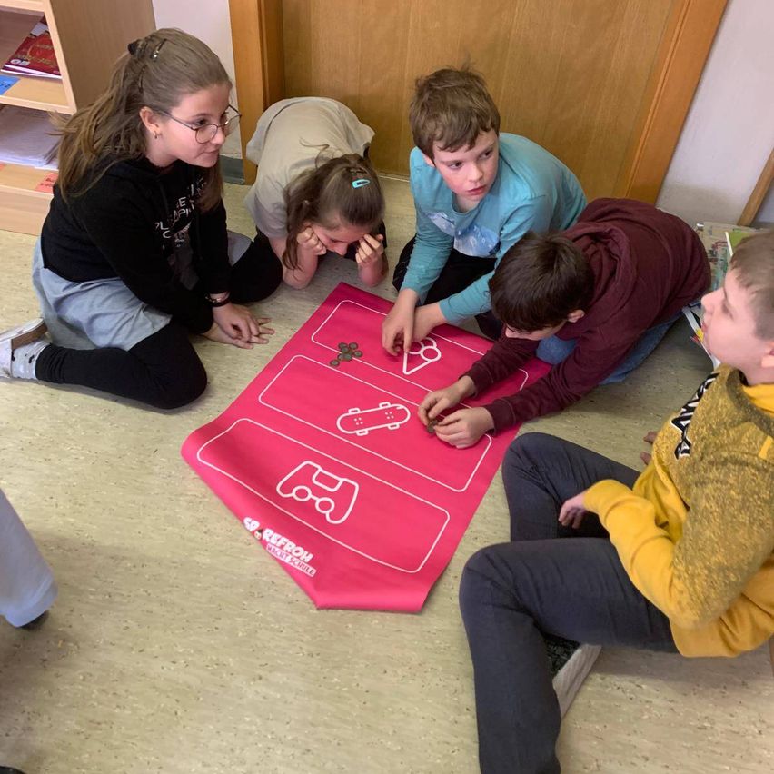 Fünf Kinder versammeln sich um eine rosa Matte mit Spielsymbolen und legen Münzen darauf. Zwei knien, drei sitzen auf dem Boden.