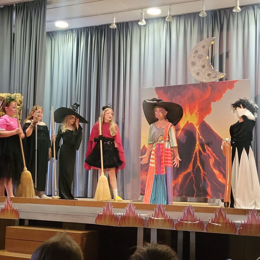 Eine Gruppe von Kindern in Hexenkostümen führt auf der Bühne mit einem Hintergrund von Vulkan und Mond auf.