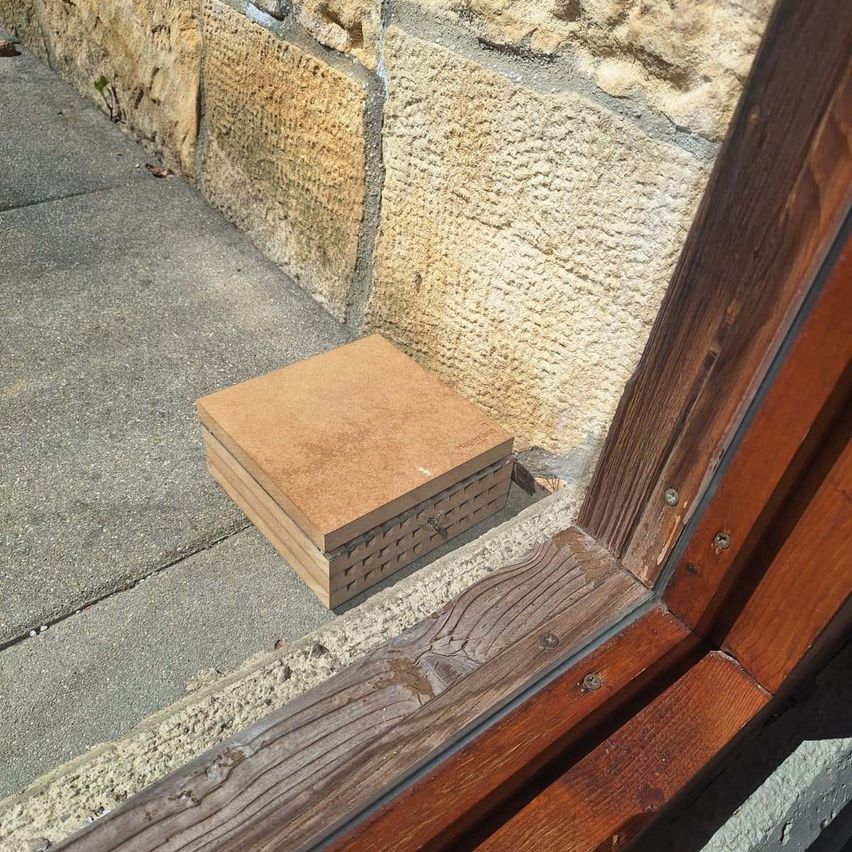 Ein kleiner Stapel brauner Holzbretter liegt auf einem Betonpflaster in der Nähe einer Steinmauer und eines Holzfensterrahmens.