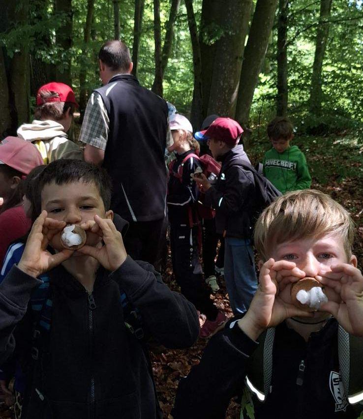 Eine Gruppe von Kindern und Erwachsenen im Wald, zwei Jungen essen Kekse, andere stehen im Hintergrund, tragen Jacken und Mützen.