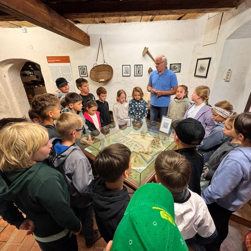 Eine Gruppe von Kindern, einschließlich Jungen und Mädchen, ist um eine Glasschaufenster in einem Museum versammelt. Ein älterer Mann steht vor ihnen und spricht. Der Raum hat Holzbalken und eine Wand mit verschiedenen Bildern und einem Schild.