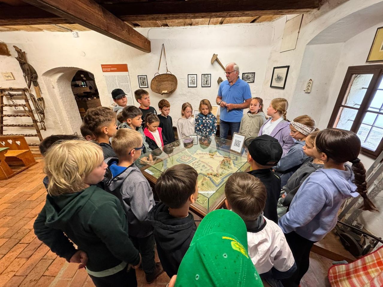 Eine Gruppe von Kindern, einschließlich Jungen und Mädchen, ist um eine Glasschaufenster in einem Museum versammelt. Ein älterer Mann steht vor ihnen und spricht. Der Raum hat Holzbalken und eine Wand mit verschiedenen Bildern und einem Schild.