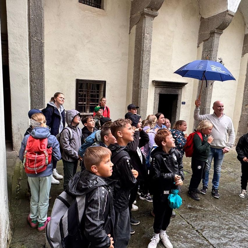 Eine Gruppe von Schulkindern und Lehrern versammelt sich vor einem Steingebäude. Einige Schüler tragen Rucksäcke. Ein erwachsener Mann hält einen Regenschirm.