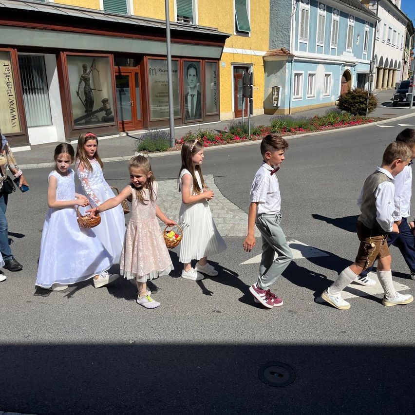 Eine Gruppe von Kindern, gekleidet in formeller Kleidung, überquert die Straße in einem Stadtplatz mit Geschäften und Blumen.