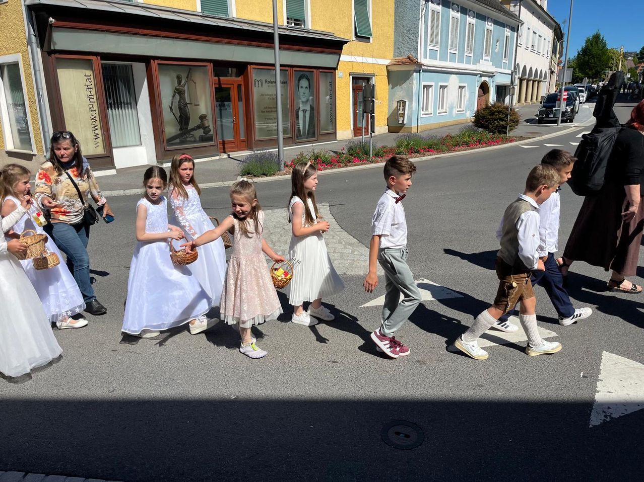 Eine Gruppe von Kindern, gekleidet in formeller Kleidung, überquert die Straße in einem Stadtplatz mit Geschäften und Blumen.