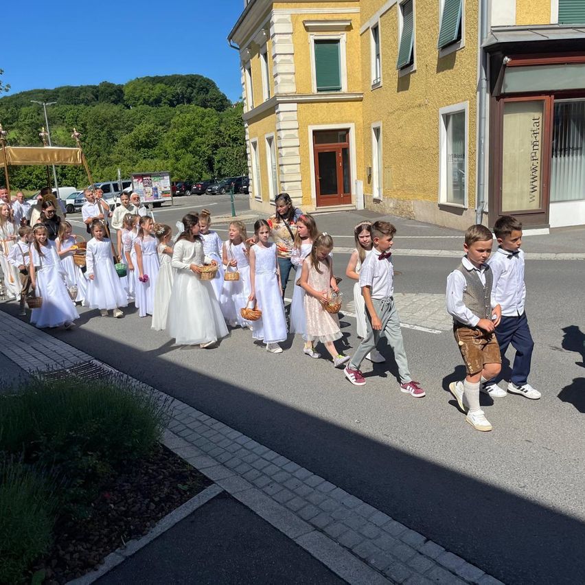 Eine Gruppe von Kindern in weißen Kleidern geht in einer Reihe, wahrscheinlich für eine religiöse Veranstaltung. Sie halten Körbe. Dahinter befindet sich ein Gebäude mit Fenstern und einer Tür.