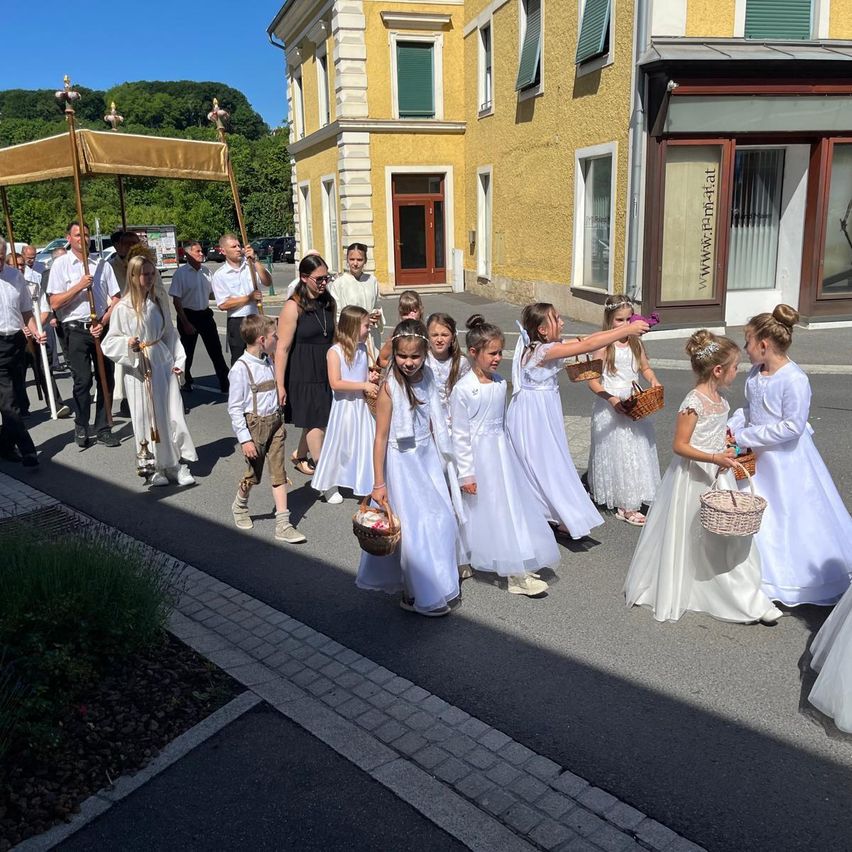 Eine Gruppe von Kindern in weißen Kleidern geht mit Körben in der Hand die Straße entlang. Dahinter stehen Erwachsene, ebenfalls in Weiß, und tragen goldene Stäbe. Gebäude säumen die Straße, eines mit dem Schild www.reha-frei.at.
