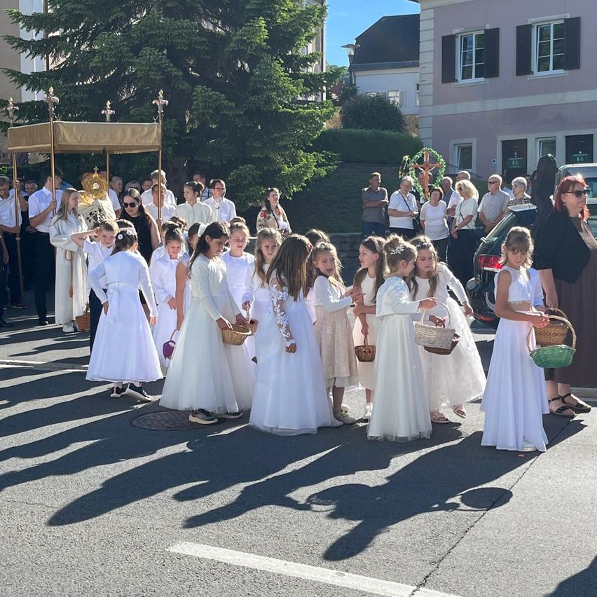 Eine Gruppe junger Mädchen in weißen Kleidern, die Körbe tragen, nimmt an einer religiösen Prozession teil, begleitet von einem Erwachsenen in einem weißen Gewand. Im Hintergrund befinden sich Gebäude, Bäume und Autos.