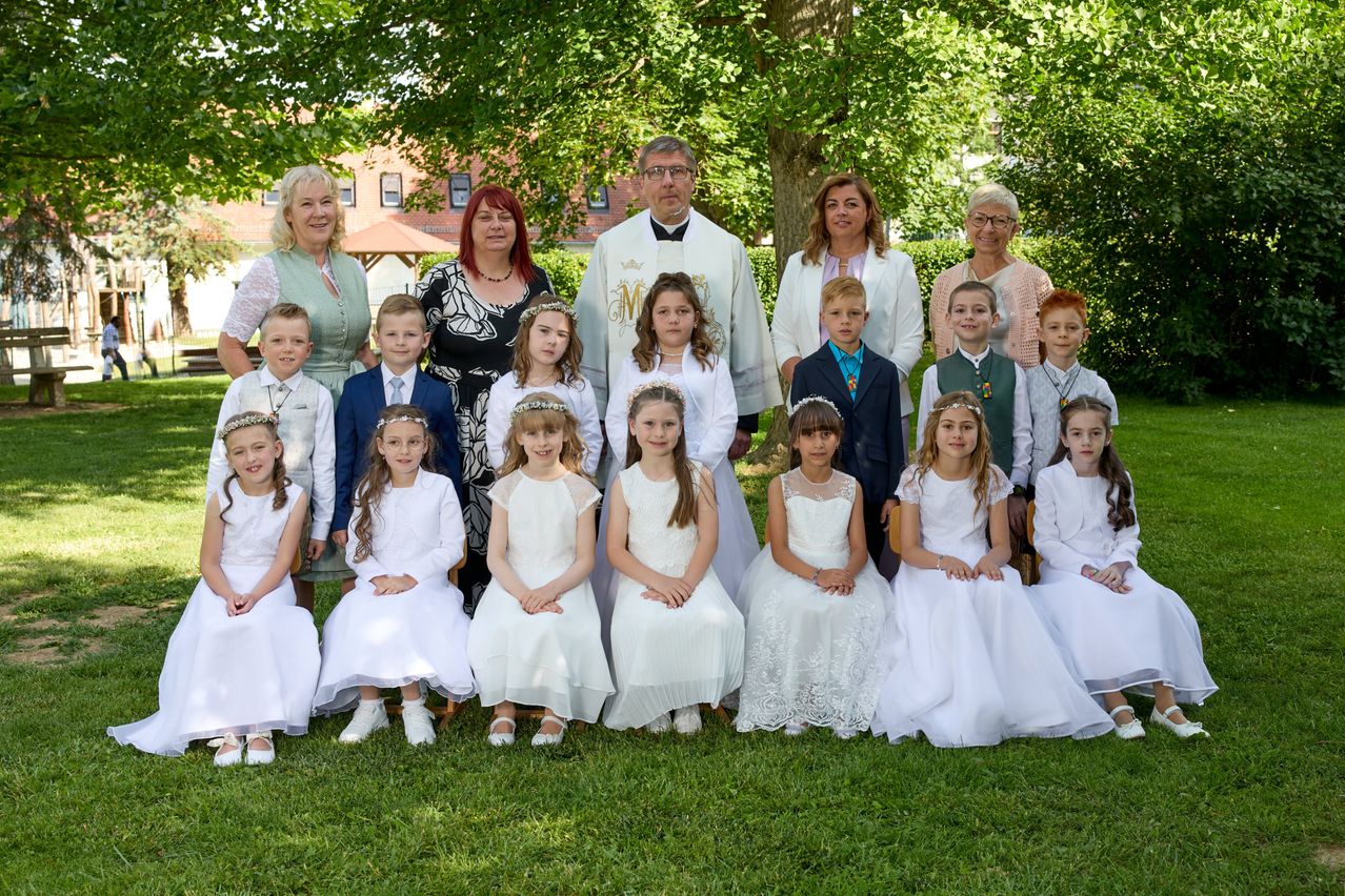 Ein Priester steht hinter einer Gruppe von Kindern und Erwachsenen in Weiß. Sie lächeln und posieren für ein Foto im Freien.