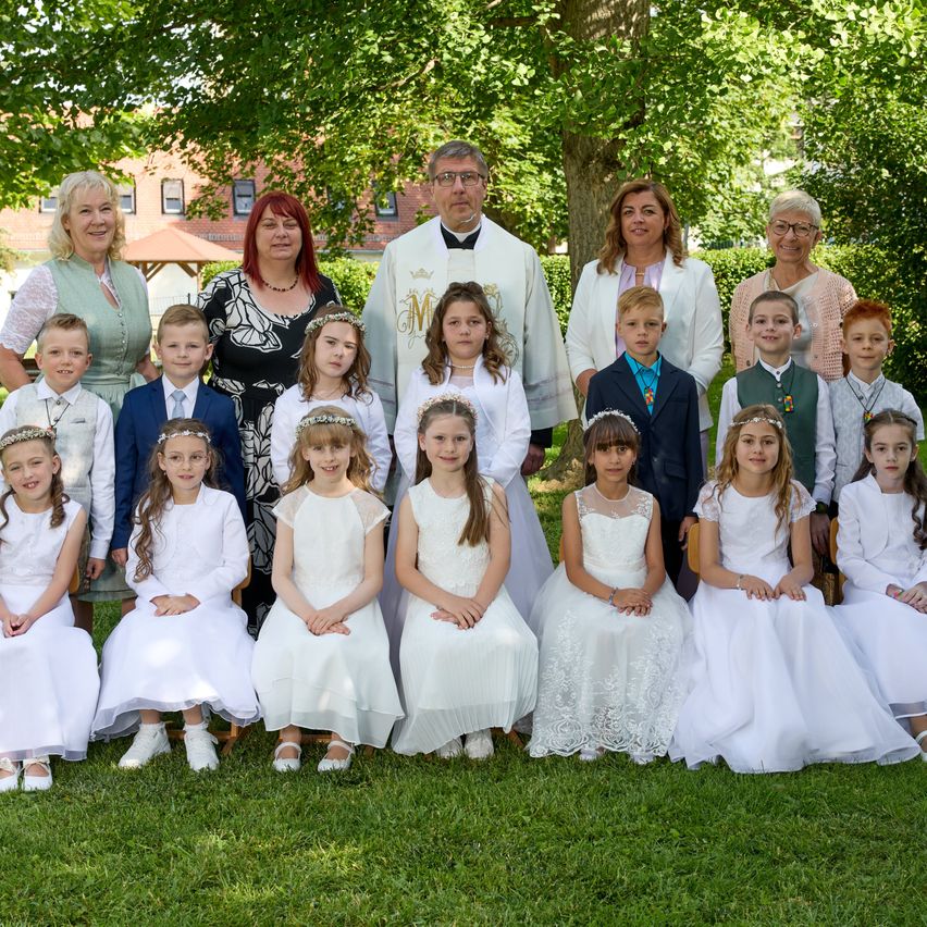 Ein Priester steht hinter einer Gruppe von Kindern und Erwachsenen in Weiß. Sie lächeln und posieren für ein Foto im Freien.