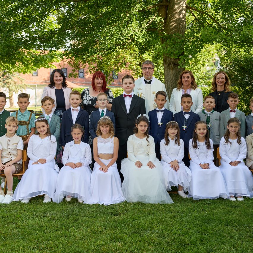 Eine Gruppe von Kindern und Erwachsenen, wahrscheinlich Familie, versammelt sich auf einem Rasen unter einem großen Baum. Sie sind alle in Weiß gekleidet.