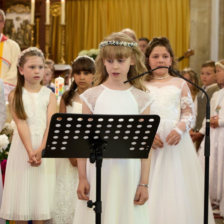 Ein junges Mädchen steht an einem Mikrofon und singt wahrscheinlich während eines Gottesdienstes, umgeben von anderen Mädchen in weißen Kleidern. Dahinter sind weitere Kinder anwesend. Im Hintergrund ist ein Priester zu sehen.