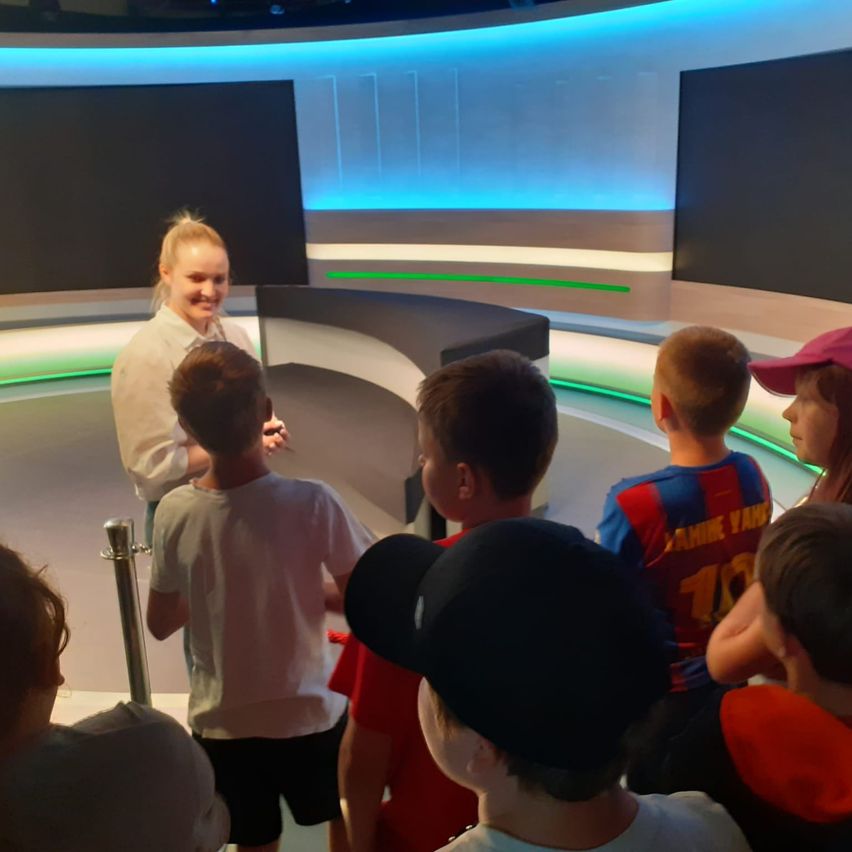 Eine Frau steht vor einer Gruppe von Kindern in einem Studio. Die Kinder tragen verschiedene Mützen, und zwei tragen Trikots. Das Studio hat blaue und grüne Lichter.